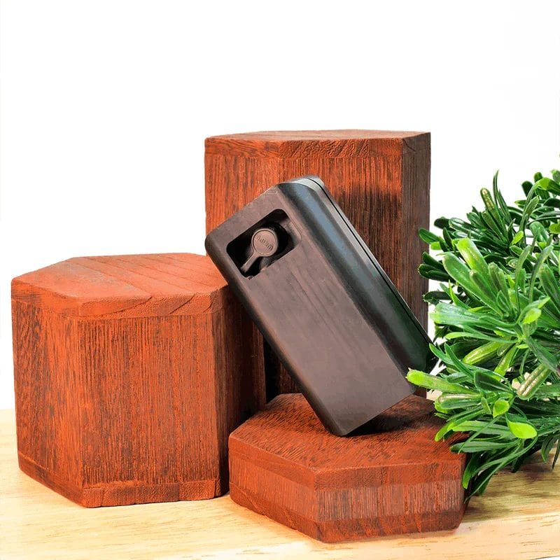 A black rectangular electronic device leaning against hexagonal wooden display blocks, accompanied by green foliage.
