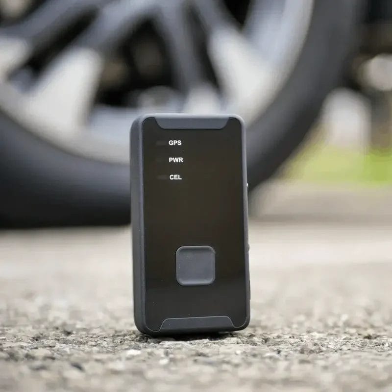 Un traceur électronique rectangulaire noir, vertical, posé sur une surface asphaltée avec un pneu de voiture flou en arrière-plan.