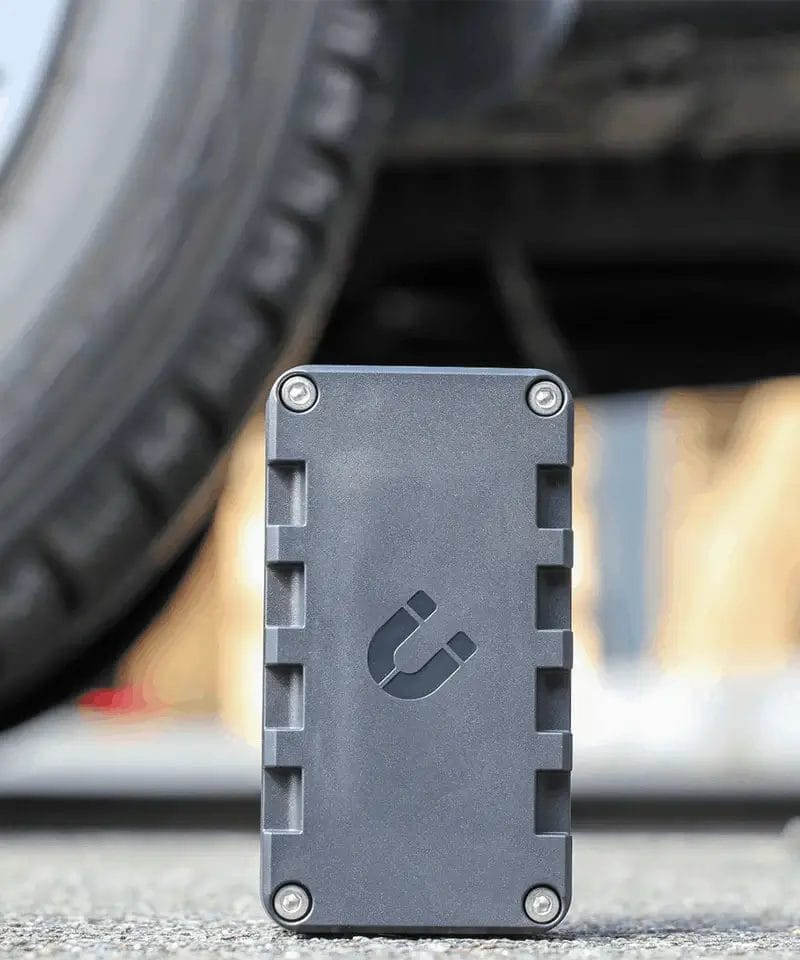 Een verticale, zwarte rechthoekige elektronische tracker die rechtop staat op een asfaltoppervlak met een wazige autoband op de achtergrond.