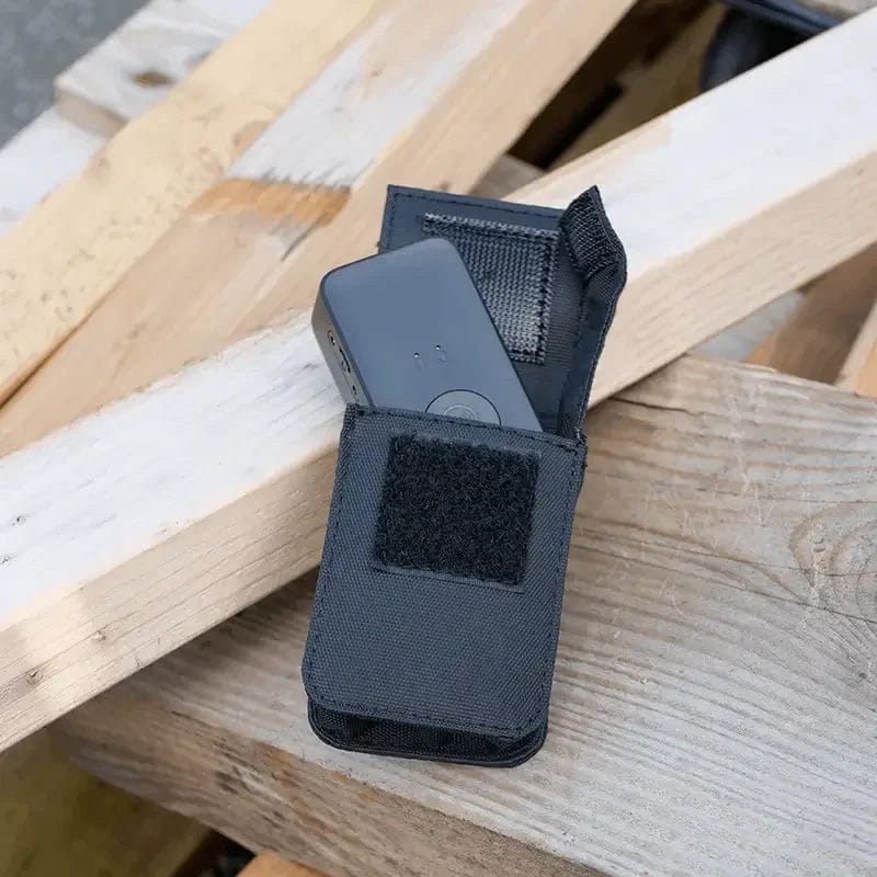 Un appareil électronique rectangulaire gris foncé partiellement glissé dans une pochette de ceinture en nylon noir ouverte, posée en diagonale sur des palettes de construction en bois empilées.